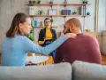 Un couple en consultation avec une psychologue conjugale. La femme pose une main réconfortante sur l'épaule de son partenaire.