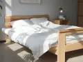 Ein helles Holzbett mit weißer Bettwäsche, das man leicht selber bauen kann. Sonnenschein fällt durch das Fenster.