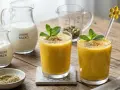 Dwa szklanki orzeźwiającego mango lassi, udekorowane miętą, na drewnianym stole. Obok składniki: pokrojone mango, jogurt, mleko, miód i przyprawy.