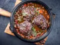 Carrilleras de ternera estofadas en salsa oscura con zanahorias y hierbas, servidas en una cazuela de hierro fundido sobre tabla de madera.