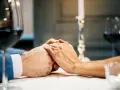 Dłonie pary splecione na stole, ozdobione złotym pierścionkiem. Romantyczna kolacja, świece i wino. To może być ich 100 rocznica ślubu.