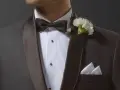 Mężczyzna w ciemnej marynarce z czarną muszką i białym kwiatem w butonierce.