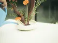 Ręka podlewa niebieską konewką kompozycję ikebana co to. Pomarańczowe gerbery, liście i gałązki z koralikami w białej misie.