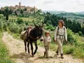 Una donna e un bambino camminano su sentieri Val d'Orcia con un asino carico, sullo sfondo un borgo medievale.