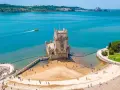 Wieża Belém, symbol kultury Portugalii, dumnie stoi nad wodą. Turyści spacerują po plaży i nabrzeżu.