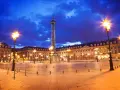 La majestuosa plaza Vendôme se ilumina bajo un cielo nocturno azul intenso, con farolas doradas y la imponente columna en el centro.
