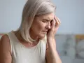 Kobieta z siwymi włosami, zamyślona, trzyma dłoń na skroni, jakby odczuwała ból głowy.