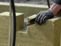Ręka w rękawicy tnie piłą wełnę skalną 10. Materiał izolacyjny jest przygotowywany do montażu.