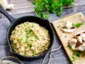 Risotto z grzybami w czarnej patelni, obok deska z pieczarkami i natką pietruszki.