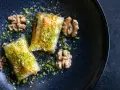 Zwei Stück Baklava, bestreut mit gehackten Pistazien und Nüssen, auf einem dunklen Teller. Ein wahrhaftiges Baklava Rezept Original.