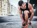 Mężczyzna w słuchawkach zawiązuje sznurówki pomarańczowych butów, przygotowując się do treningu. W tle miejski krajobraz z budynkami. Ćwiczenia takie jak nordic curl wzmocnią jego nogi.