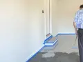 Mann beschichtet den garagenboden mit einem Roller. Der Boden ist teilweise mit einer grauen Beschichtung bedeckt, die Ränder sind mit blauer Klebefolie abgeklebt.
