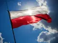Biało-czerwona flaga Polski powiewa na tle błękitnego nieba z chmurami. Słońce przebija się przez materiał, podkreślając jego fakturę. Wymiary flagi polski są standardowe, proporcje zachowane.