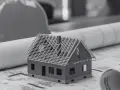 Model domu na planach budowlanych, z widoczną szerokością elewacji frontowej. Obok kask budowlany i zwinięte projekty.
