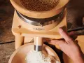 Holzgetreidemühle mit Getreide oben und frisch gemahlenem Mehl unten, Hand bedient die Mühle.