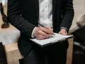 Prawnik w czarnym garniturze podpisuje pozew o zapłatę na dokumencie. W tle widać figurkę Temidy.