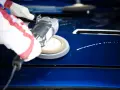 A gloved hand uses a buffer to polish a shiny blue car hood, demonstrating how to polish a car with a buffer for a flawless finish.