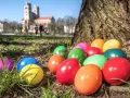 Bunte Ostereier liegen im Gras vor einem Baum. Im Hintergrund ist eine Kirche zu sehen. Die Suche nach den Eiern ist ein schöner Teil der Ostern Bedeutung.