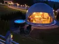 Nocny widok na luksusowy camping w Tatrach. Dwa kopułowe namioty, jeden z wanną i łóżkiem, drugi z oświetleniem. Obok jacuzzi i meble ogrodowe.