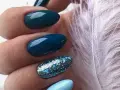 Unghie per l'inverno: unghie blu petrolio e azzurre con glitter scintillanti, accanto a una soffice piuma bianca.