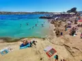 Słoneczna plaża na Malcie w lutym. Ludzie kąpią się w turkusowej wodzie, opalają na piasku i odpoczywają pod parasolami.