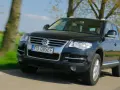 Czarny VW Touareg 3.0 TDI w ruchu, z widocznymi wadami lakieru i drobnymi niedoskonałościami, na tle zielonych pól i drzew.
