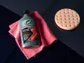 En flaska Turtle Wax Hybrid Solutions Ceramic Polish & Wax, en rosa mikrofiberduk och en polertrissa ligger på en mörkblå yta.