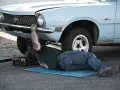 Un uomo lavora sotto un'auto d'epoca, forse per un intervento di **art 46 C.d.S.**, con un cric che la solleva.