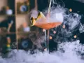 Kieliszek z drinkiem otoczony gęstym dymem do drinków, z pomarańczową skórką jako dekoracją.