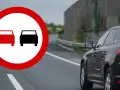 Znak zakazu wyprzedzania na drodze. Czarny SUV jedzie po prawej stronie, a z lewej widać znak z dwoma samochodami.