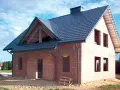 Budowa domu z cegły, widoczny dach betonowy skośny ocieplenie. Widać otwory okienne i drzwiowe, komin.