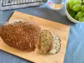 Chleb z twarogu, bogaty w ziarna i orzechy, pokrojony na desce. Obok miska z winogronami i szklanka wody.