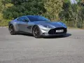Srebrny Aston Martin Vanquish 2025 z brązowymi felgami na tle zieleni.