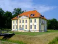 Pałac w Choroszczy, żółty, z pomarańczowym dachem, otoczony zielenią. Drewniany mostek prowadzi do posiadłości.