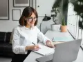 Uśmiechnięta kobieta w okularach pisze w notatniku, pracując przy laptopie. Zastanawia się, ile zarabia redaktor.