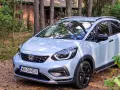 Jasny Honda Jazz Crosstar zaparkowany w lesie, obok drewnianego domku.