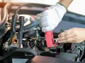 Eine Hand mit Handschuh klemmt das rote Kabel an den Pluspol, während die andere Hand den Minuspol der Autobatterie sichert.