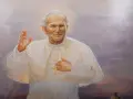 Obraz przedstawia Jana Pawła II w białej szacie, z podniesioną prawą dłonią i krzyżem na piersi. Wierni odmawiają modlitwę do Jana Pawła II o uzdrowienie.