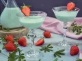 Zwei Gläser mit hellgrünem Likör, garniert mit Erdbeeren. Ein Krug mit dem Likör und frische Erdbeeren deuten darauf hin, dass man Waldmeister Schnaps selber machen kann.