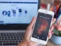 Eine Hand hält ein iPhone mit gesperrtem Bildschirm und Ziffernfeld. Im Hintergrund ein Laptop.