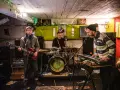 Un groupe de musique punk des années 90 joue dans un local sombre. Un guitariste, un batteur et un claviériste sont sur scène.