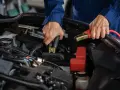 Hände mit Starthilfekabeln, um die **Batterie richtig anzuklemmen**. Ein Mechaniker in blauer Arbeitskleidung verbindet die Klemmen mit der Autobatterie.