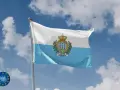 Flaga San Marino powiewa na tle błękitnego nieba z chmurami. Herb z trzema wieżami i koroną zdobi biało-błękitny materiał.
