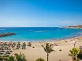 Spiaggia affollata con ombrelloni e lettini, mare azzurro e città costiera sullo sfondo. Sembra l'isola più bella delle Canarie.