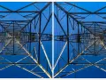 Widok z dołu na kratownicę słupa energetycznego, tworzącą geometryczny wzór na tle błękitnego nieba. To fotografia stereoskopowa.
