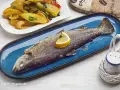 Eine ganze Forelle im Backofen, garniert mit einer Zitronenspalte, liegt auf einem blauen Teller. Daneben eine Beilage aus Kartoffeln und Gemüse.