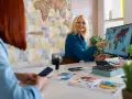 Kobieta wskazuje na mapę świata na monitorze komputera, siedząc przy biurku z materiałami podróżniczymi.