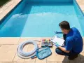 A man in blue checks pool water test tubes, preparing to shock the pool. Pool cleaning supplies are laid out.