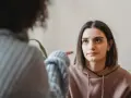 Dwie kobiety rozmawiają. Jedna wskazuje palcem na drugą, która patrzy z wyrzutem, jakby miała potworne wyrzuty sumienia.