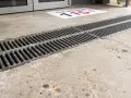 Un panneau au sol indique "Sip iPhone 115€". Un caniveau en métal longe un sol en béton. Le béton drainant prix m2 est une solution pour évacuer l'eau.